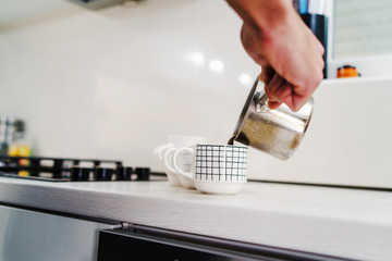 Close up on male hand caucasian holding a metal shiny pot pouring homemade black coffee in the cup on the workbench at kitchen at home in day