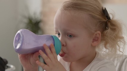 Close-up shot of fair-haired toddler girl with pigtails drinking milk from baby bottle at home