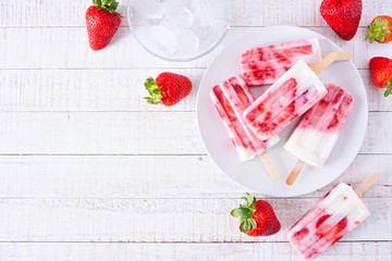 Plate of homemade strawberry vanilla yogurt popsicles. Above view corner border on a white wood background with copy space.