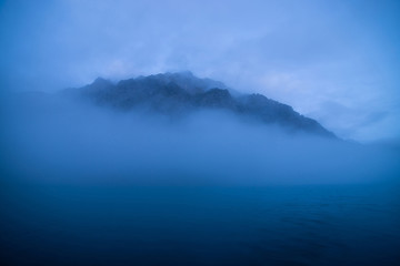 Tranquil minimal landscape with big rocky mountain in middle of water among low clouds in twilight. Wavy sea of blue classic color. Atmospheric scenery with deep blue calm lake and rock in dense fog.