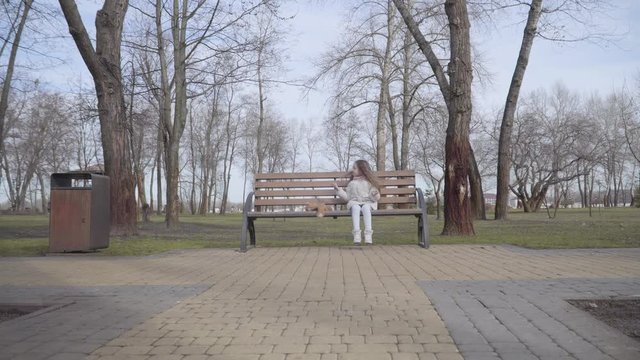 Extreme wide shot of lonely little girl sitting on bench in park. Caucasian child with long curly hair get lost in spring or autumn day. Social issues, kidnapping, loneliness, abandonment, fear.