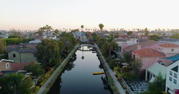Aerial, reverse, drone shot rising over a canoe on a still Canal, in between colorful houses, with Venice beach in the background, during dusk, in Los Angeles, CA, United States