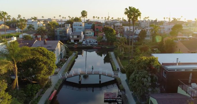 Aerial, drone shot tracking over a tranquil water channel and a man with a kid on his shoulders, walking over the canal bridge, during the Covid19 Pandemic, at sunset, in LA, CA, USA