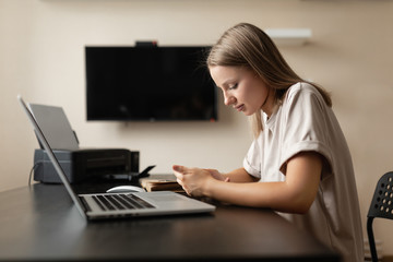 A cute pensive girl does work from home, scans information on the phone and checks it against information on the computer during quarantine and pandemic.