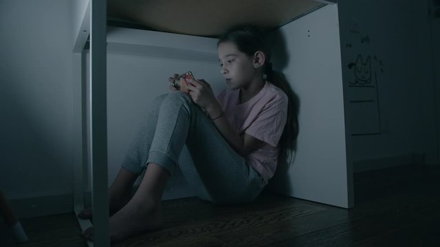Little girl emotionally plays in smartphone under table at night