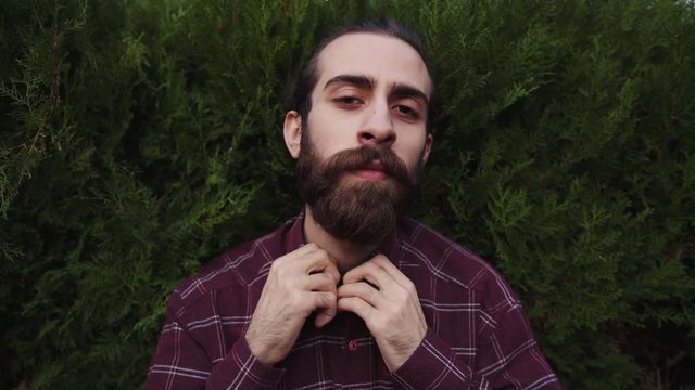 Handsome hipster young bearded man with long hair unbuttons his plaid shirt. Attractive seductive movements.