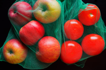 Bright red apples and tomatoes in green eco bags close up on a dark table