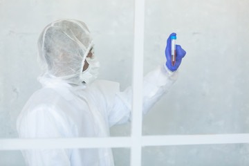 Doctor in chemical protection suit is standing behind the window and holding blood sample fot testing it on COVID, COVID-19, coronavirus to stop pandemic and epidemic of the virus