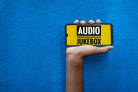 Audio Jukebox Word On Smart Phone Screen Isolated On Blue Background With Copy Space For Text. Person Holding Mobile On His Hand And Showing Front Of The Screen Wording Audio Jukebox.