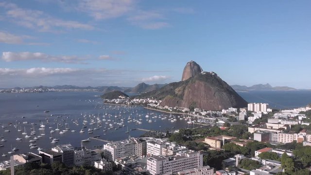 Sugarloaf mountain in the Guanabara bay with pleasure boats in the cove and Pasmado hill with viewpoint in the foreground against a blue sky with clouds. Aerial ascend revealing foreground hilltop