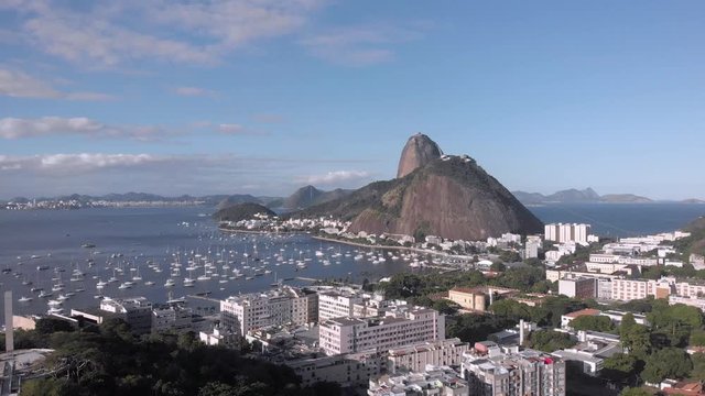 Sugarloaf mountain in the Guanabara bay with pleasure boats in the cove and Pasmado hill with viewpoint in the foreground against a blue sky with clouds. Aerial descend revealing foreground hilltop