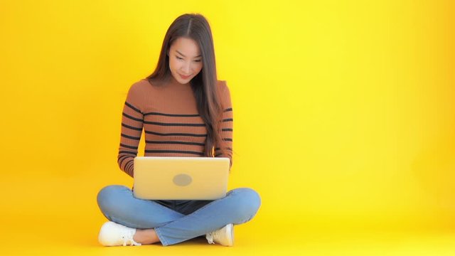 Young woman in jeans and sweater sitting with legs crossed using laptop computer smiles. Asian lady with long hair smiles while typing, internet shopping or surfing web. Bright yellow background.