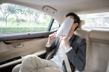Stressed young asian businessman looking at Financial documents with expression Tired and worried while sitting on the back seat in the car.  many bad financial report.