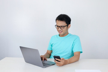 Asian young man happy and smile when he see on smart phone with laptop beside it. Indonesian man wearing blue shirt.