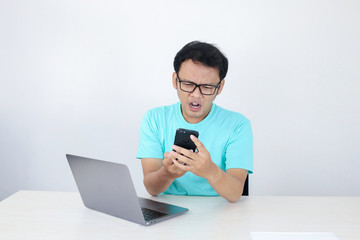 Wow face of Young Asian man shocked what he see in phone with laptop beside it. Indonesian man wearing blue shirt.