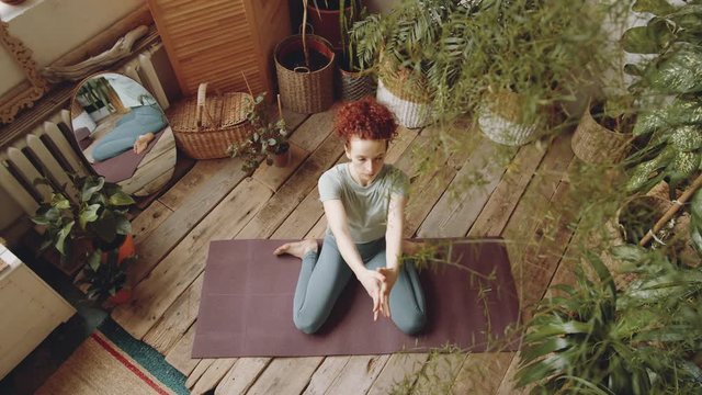 Top view of redhead woman in sports legging and t-shirt sitting in hero pose on mat and stretching her arms up while practicing yoga at home in room with lots of green plants and wooden floor