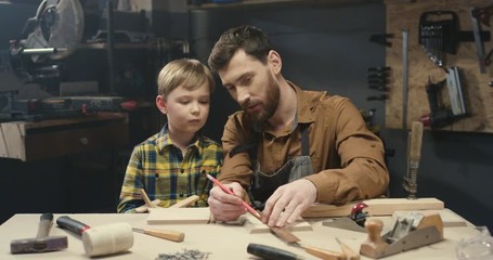 Handsome young Caucasian father carpenter teaching how to work with wood his small cute son in workshop. Woodworker showing to little boy hardwood proceeding and furniture producing. - Powered by Adobe