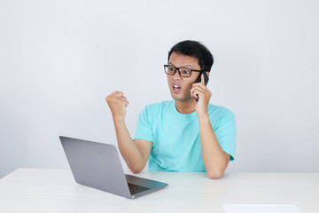 Wow face of Young Asian man shocked what he see in laptop when calling on phone. Indonesian man wearing blue shirt.