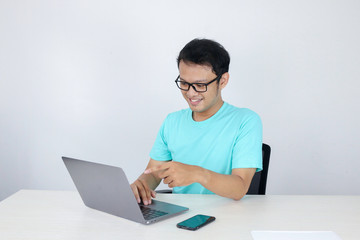 Young Asian man feeling happy and smile when work laptop on table. Indonesian man wearing blue shirt.