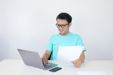 Young Asian Man is smile and happy when working on a laptop and document on hand. Indonesian man wearing blue shirt.