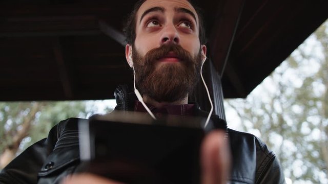 Young handsome bearded man is texting,listening music on phone with earphones and smiling. He is sitting in park outdoors.