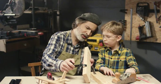 Caucasian senior man carpenter teaching his small cute grandson how to work with wood in workshop at desk. Little boy watching how his grandfather woodman working with hardwood.