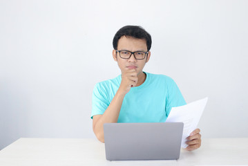 Young Asian Man is serious and focus when working on a laptop and document on the table. Indonesian man wearing blue shirt.