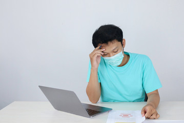 Young Asian man wearing medical mask is feeling unhealthy, tired, and confused with working in documents and laptops on the table.