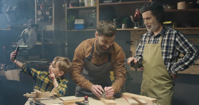 Cute little boy taking selfie photos with smartphone camera in carpentry workshop while his father proceeding wood and grandfather smoking and resting. Family of carpenters. Woodman with son and dad.