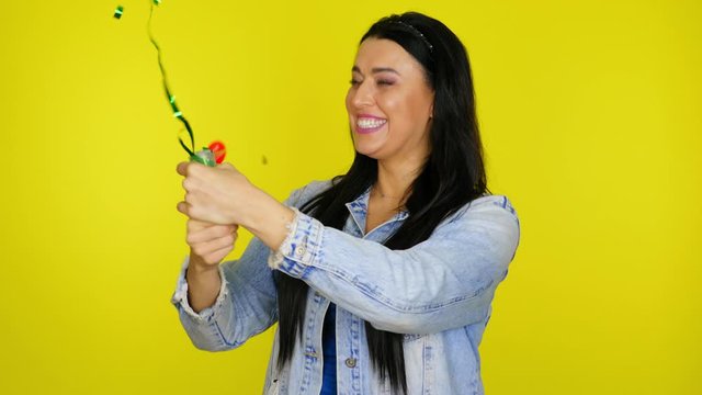 Cheerful woman with black hair in a denim jacket explodes a confetti cannon on a yellow background with copy space. Place for text or product. 4K footage