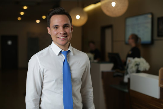 A young hotel administrator in a white shirt and blue tie stands against the front desk. Looks at the camera and smiles