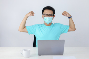 Fototapeta na wymiar Young Asian Man wearing medical mask is give empower sign with strong hand when working on a laptop on the table. Indonesian man wearing blue shirt.