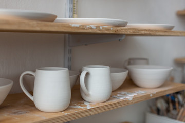 Naklejka na ściany i meble Clay blanks of handmade dishes for painting, plates and mugs of different sizes. On the shelf in the pottery workshop