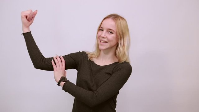Young Blonde Woman In Black Sweater With Stylish Watch On White Background, Confident Girl Shows Arm Muscles, Demonstrates Biceps. The Concept Of Strong People