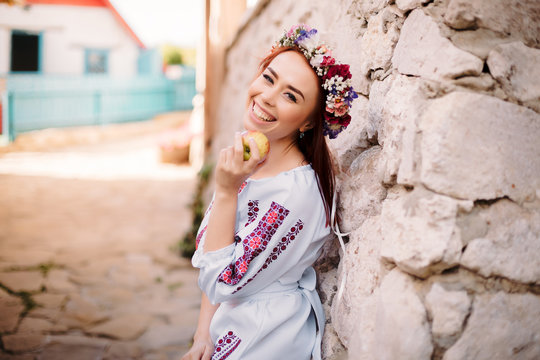 Young redhead moldavian girl dressed in traditional costume with flower wreath on the head standing near old wall.