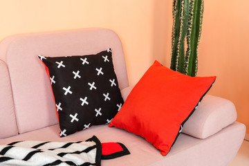 Two double-side pillows (red and black with white crosses) and same style plush plaid lying on the pink sofa in a living room with peach walls and a cactus.