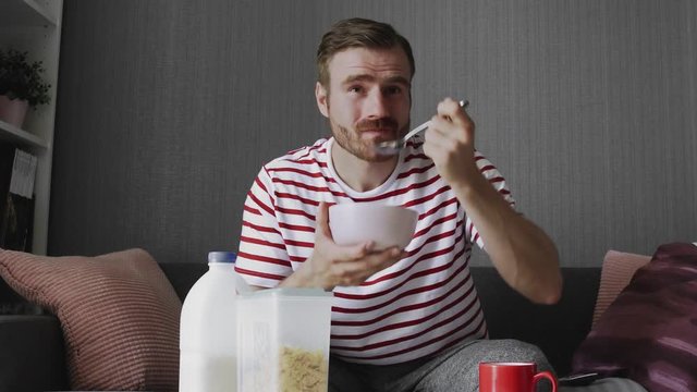 Young man watching funny TV show during breakfast sitting on sofa in room