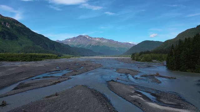 A broad riverbed of a river in Alaska. On the bottom of the mountains is forest and the peaks are covered a bit in the snow.