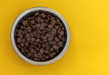 Dry brown pet food for cats or dogs in a metal bowl on a yellow background top view mock up, flat lay. Space for text
