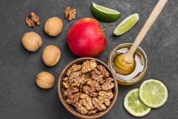 Walnut kernels in wooden bowl, honey in glass jar, lime slices and red apple on table.