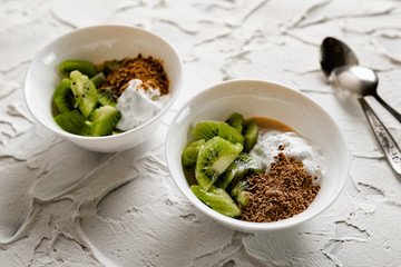 A pair of small dessert bowls with fresh kiwi fruit, white ice cream, yogurt and chocolate chips on a white stone background with teaspoons