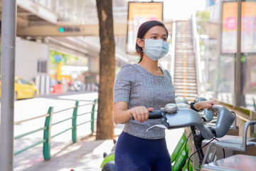 Young Asian woman with mask riding bike at public bicycle service station