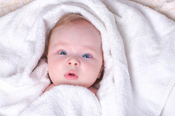Charming, surprised, blue-eyed baby with plump lips and blond hair lies in dry, white towel after bathing in bathroom. Concept of skin care, child hygiene, washing and happy childhood