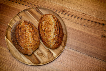 Freshly made grain sourdough bread on a wooden plater viewed from the top. The two loafs are golden and crusty.