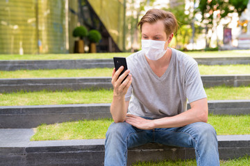 Fototapeta premium Young man with mask for protection from corona virus outbreak using phone while sitting outdoors