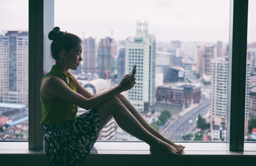 Woman using mobile phone relaxing by condo window at home or office room silhouette. Business woman...