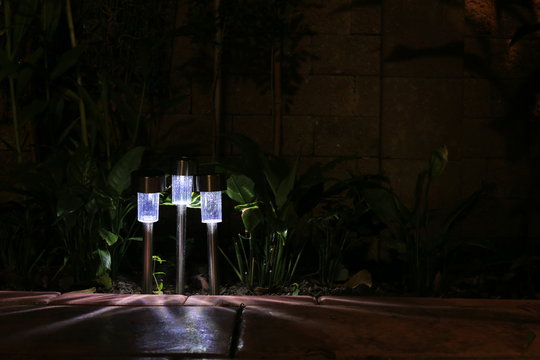 Garden LED Lamp Lighting. Small Solar Lighting Shining In A Garden And Casting Shadow In Multiple Direction. Cool Light Temperature For Modern Hi Tech Landscape Design.