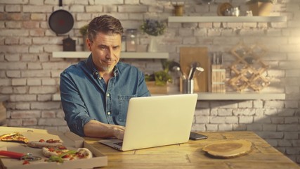 Man working from home on laptop computer, sitting at table in kitchen, eating online ordered pizza.