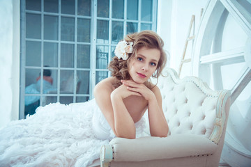 Fototapeta na wymiar Lovely young girl bride lies on a sofa in a bright studio. wedding photo shoot. Wedding hairstyle. Fashion photography in a chic white long puffy dress