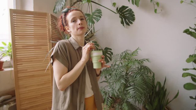 Arc Shot Of Young Positive Woman Drinking From Reusable Eco-friendly Cup With Glass Straw And Then Looking At Camera With Smile While Standing In Beautiful Room With Green Houseplants